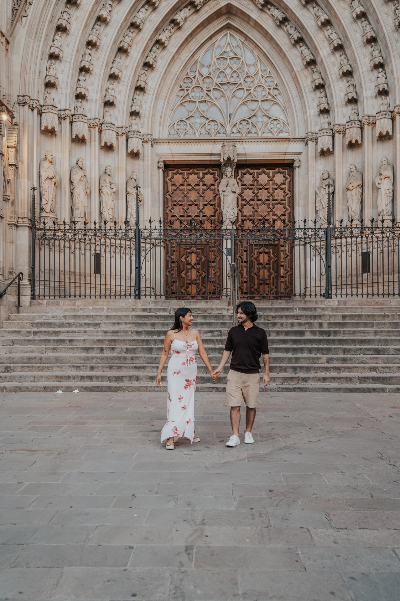 The couple holding hands in front of Barcelona Cathedral’s carved entrance and broad stone steps during their couple photoshoot in Barcelona.