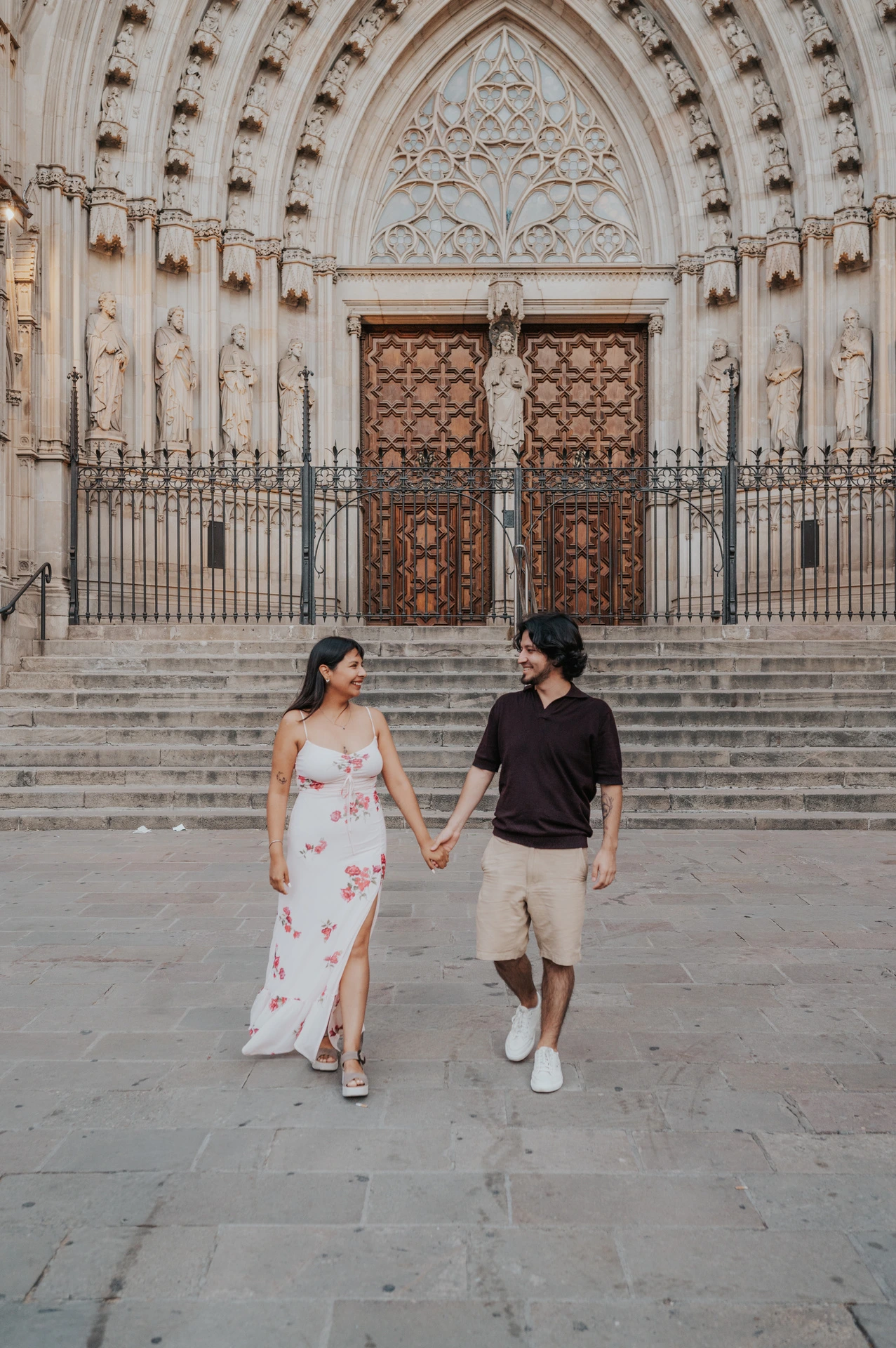 The couple walking towards the camera and looking at each other in front of Barcelona Cathedral’s carved entrance during their couple photoshoot in Barcelona.
