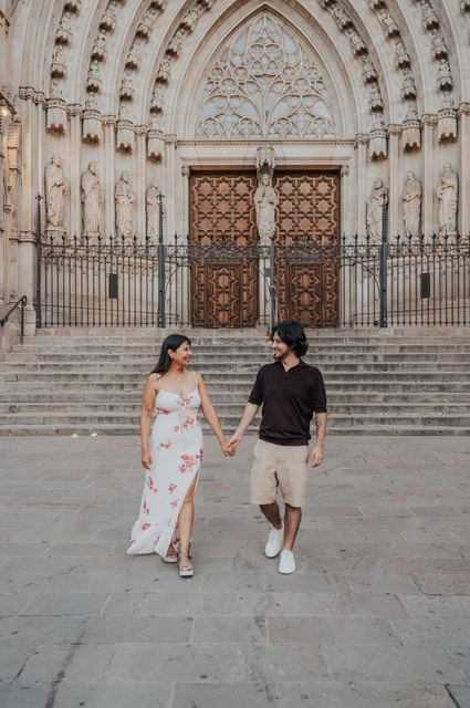 The couple walking towards the camera and looking at each other in front of Barcelona Cathedral’s carved entrance during their couple photoshoot in Barcelona.