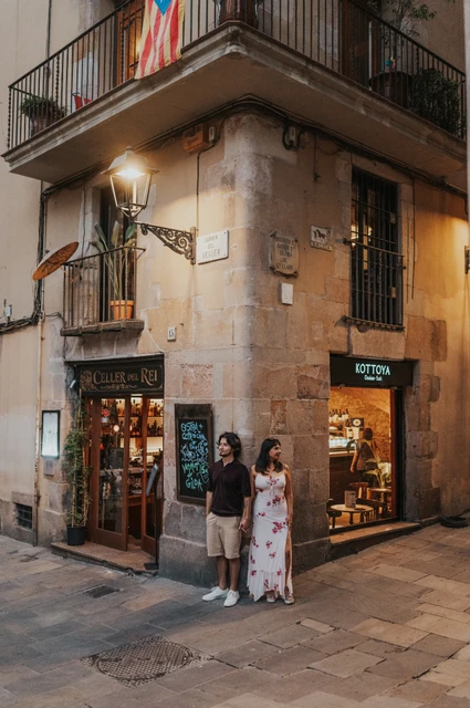 A portrait beside a glowing old-town shop corner | Barcelona couple photoshoot The couple standing together beside a corner shop beneath a glowing wall lamp in the Gothic Quarter during their couple photoshoot in Barcelona.