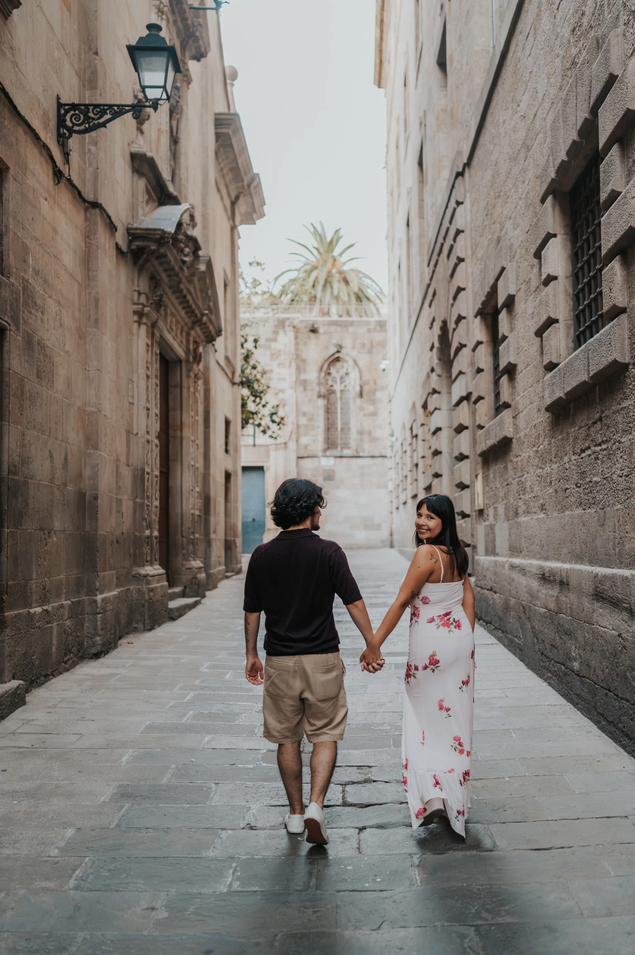 The couple walking away hand in hand along a narrow Gothic Quarter lane, with the woman looking back during their couple photoshoot in Barcelona.