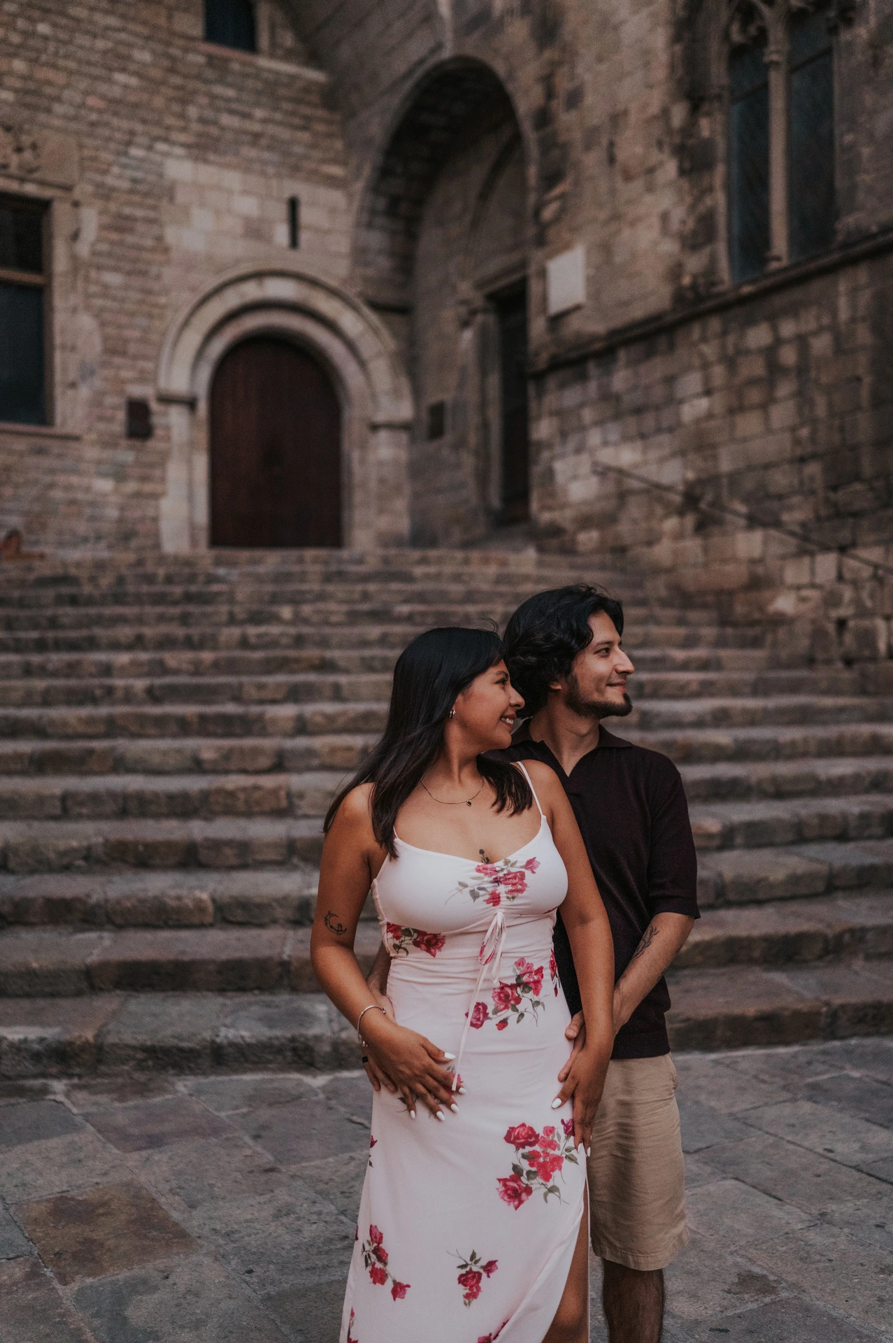 The couple standing close together and looking to one side, with the broad stone steps at Plaça del Rei behind them during their couple photoshoot in Barcelona.