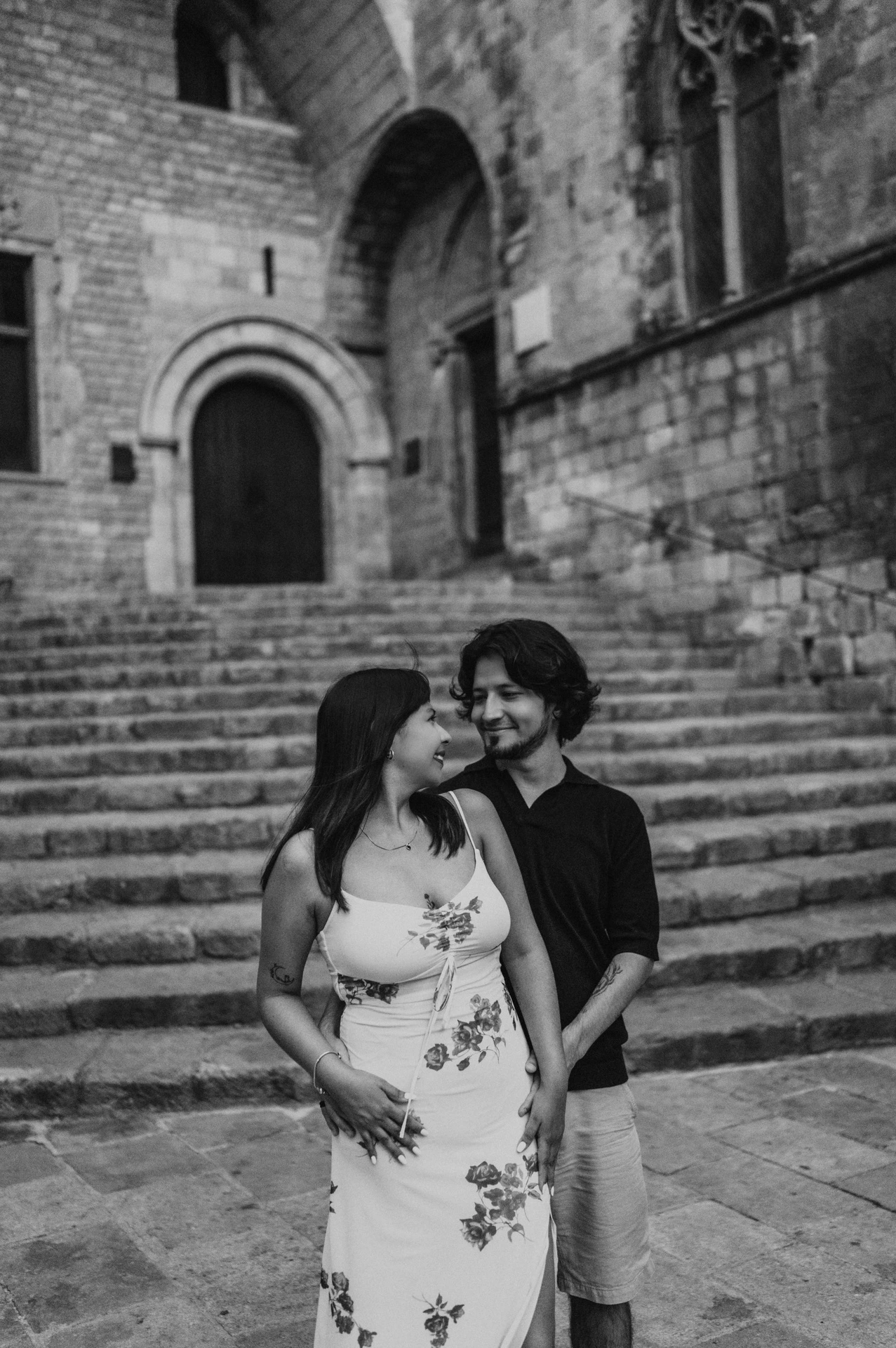 Black-and-white photograph: a close portrait of the couple smiling at each other, with the stone steps at Plaça del Rei behind them during their couple photoshoot in Barcelona.
