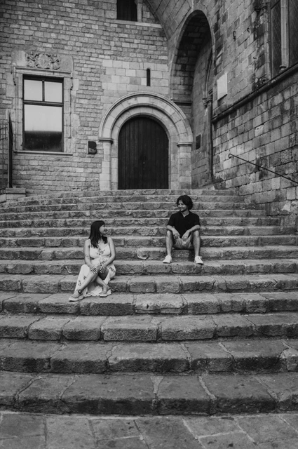 Black-and-white photograph: the couple sitting on different levels of the broad stone steps at Plaça del Rei and looking at each other during their couple photoshoot in Barcelona.