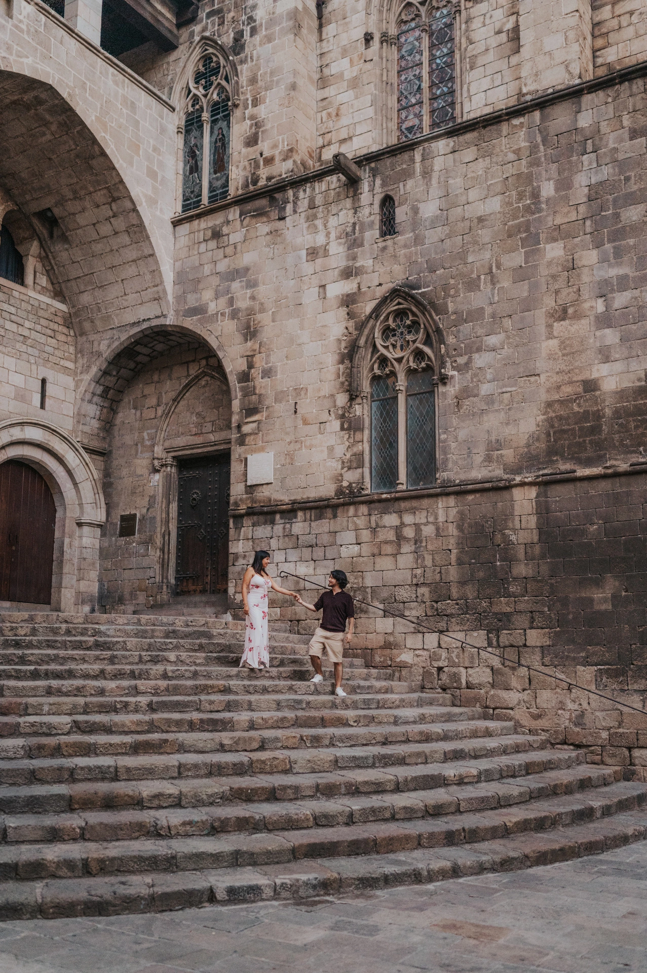 A wide view of the couple facing one another on the stone steps at Plaça del Rei, surrounded by tall stone walls and Gothic windows during their couple photoshoot in Barcelona.