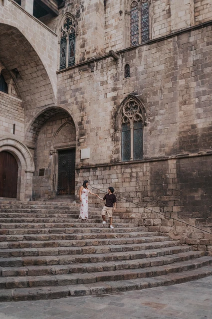 The stone steps and Gothic windows of Plaça del Rei | Barcelona couple photoshoot A wide view of the couple facing one another on the stone steps at Plaça del Rei, surrounded by tall stone walls and Gothic windows during their couple photoshoot in Barcelona.