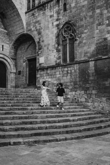 Hand in hand on the steps of Plaça del Rei in black and white | Barcelona couple photoshoot Black-and-white photograph: the couple holding hands on the stone steps at Plaça del Rei, with a tall arched doorway and Gothic windows behind them during their couple photoshoot in Barcelona.