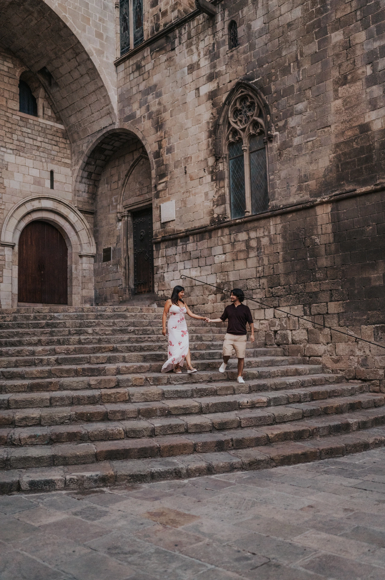 The couple holding hands on the stone steps at Plaça del Rei, with a tall arched doorway and Gothic windows behind them during their couple photoshoot in Barcelona.