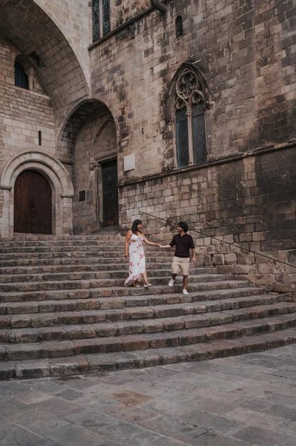 Hand in hand on the steps of Plaça del Rei | Barcelona couple photoshoot The couple holding hands on the stone steps at Plaça del Rei, with a tall arched doorway and Gothic windows behind them during their couple photoshoot in Barcelona.