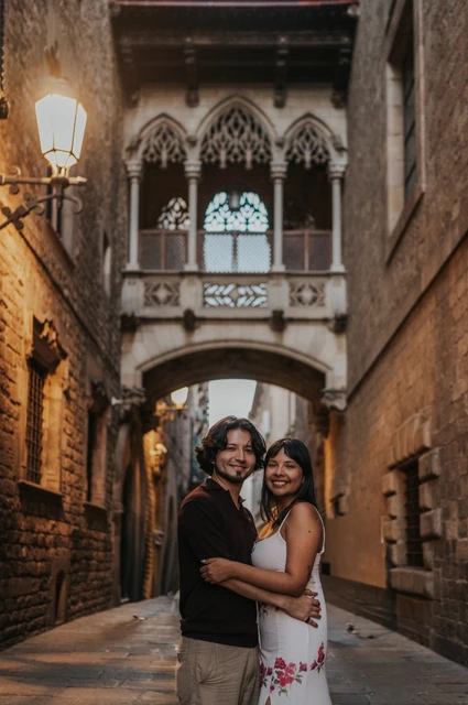 A close portrait beneath Pont del Bisbe | Barcelona couple photoshoot A close portrait of the couple smiling at the camera beneath Pont del Bisbe and a glowing wall lamp during their couple photoshoot in Barcelona.