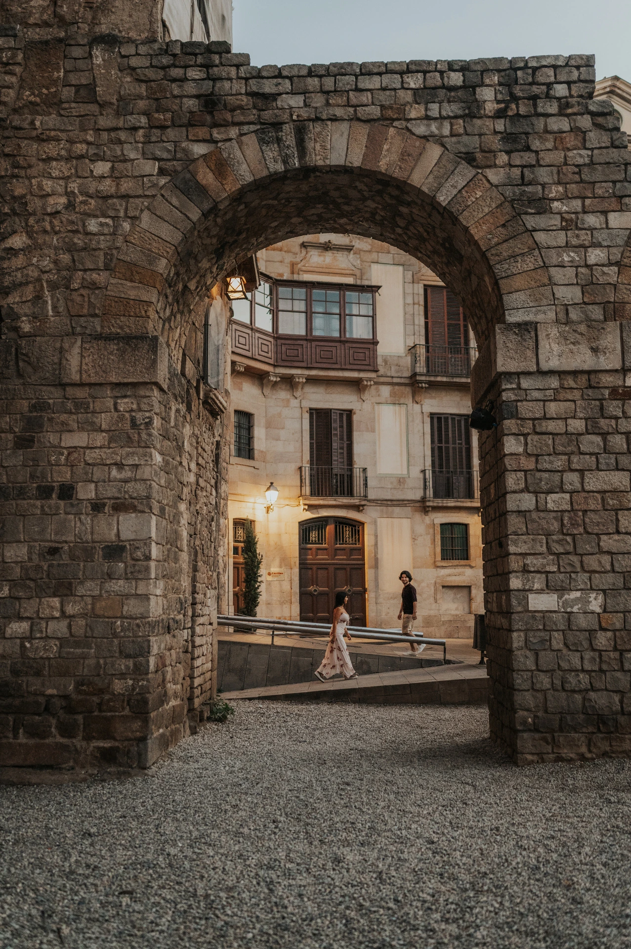 The couple walking hand in hand beyond a broad stone arch in the Gothic Quarter, with lit lamps in the street behind them during their couple photoshoot in Barcelona.