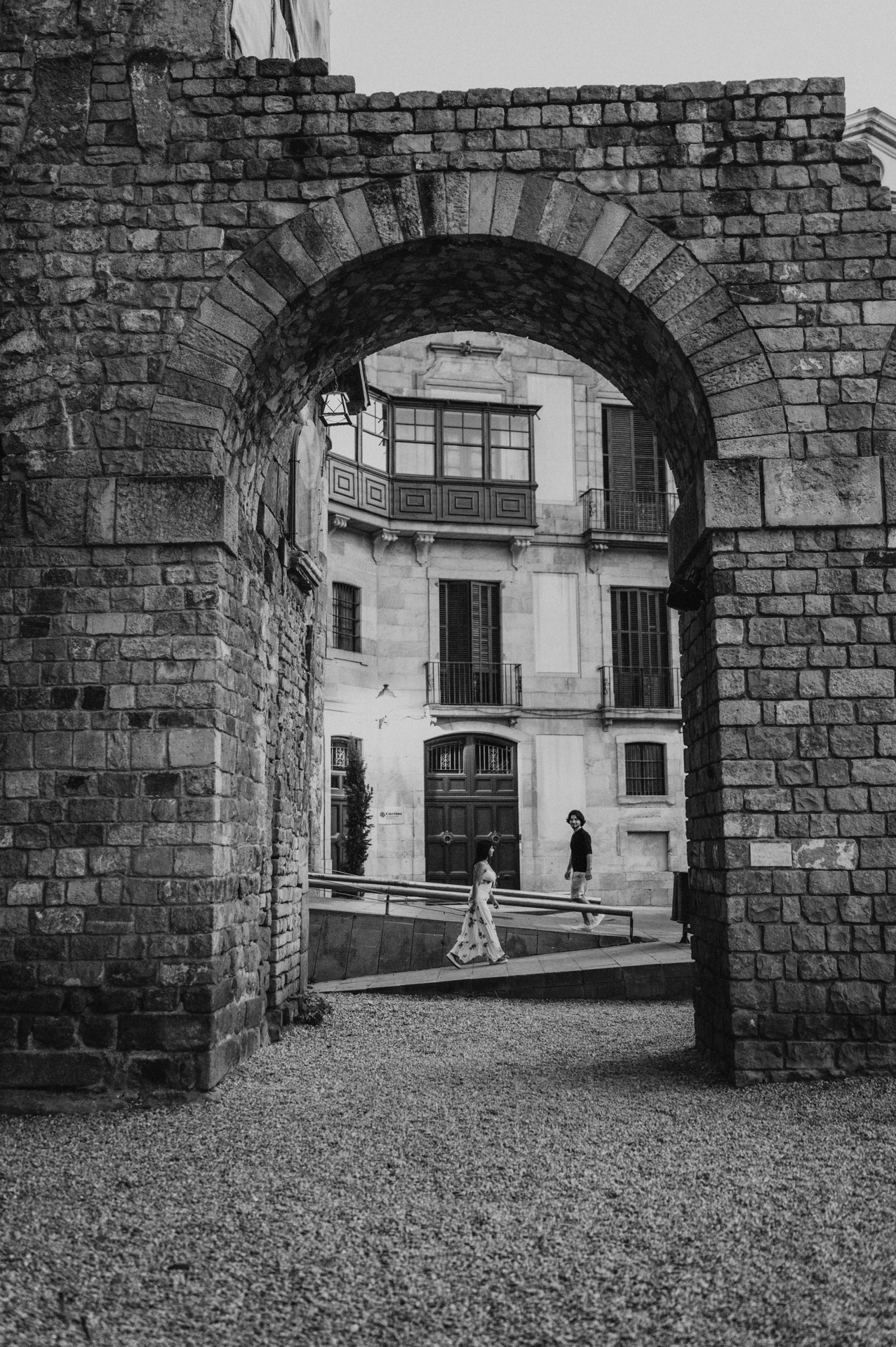 Black-and-white photograph: the couple walking hand in hand beyond a broad stone arch in the Gothic Quarter, with lit lamps in the street behind them during their couple photoshoot in Barcelona.
