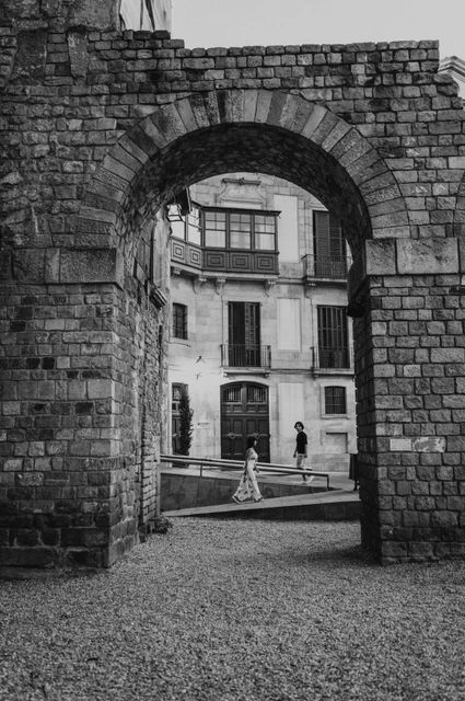 Through a stone arch in the Gothic Quarter in black and white | Barcelona couple photoshoot Black-and-white photograph: the couple walking hand in hand beyond a broad stone arch in the Gothic Quarter, with lit lamps in the street behind them during their couple photoshoot in Barcelona.