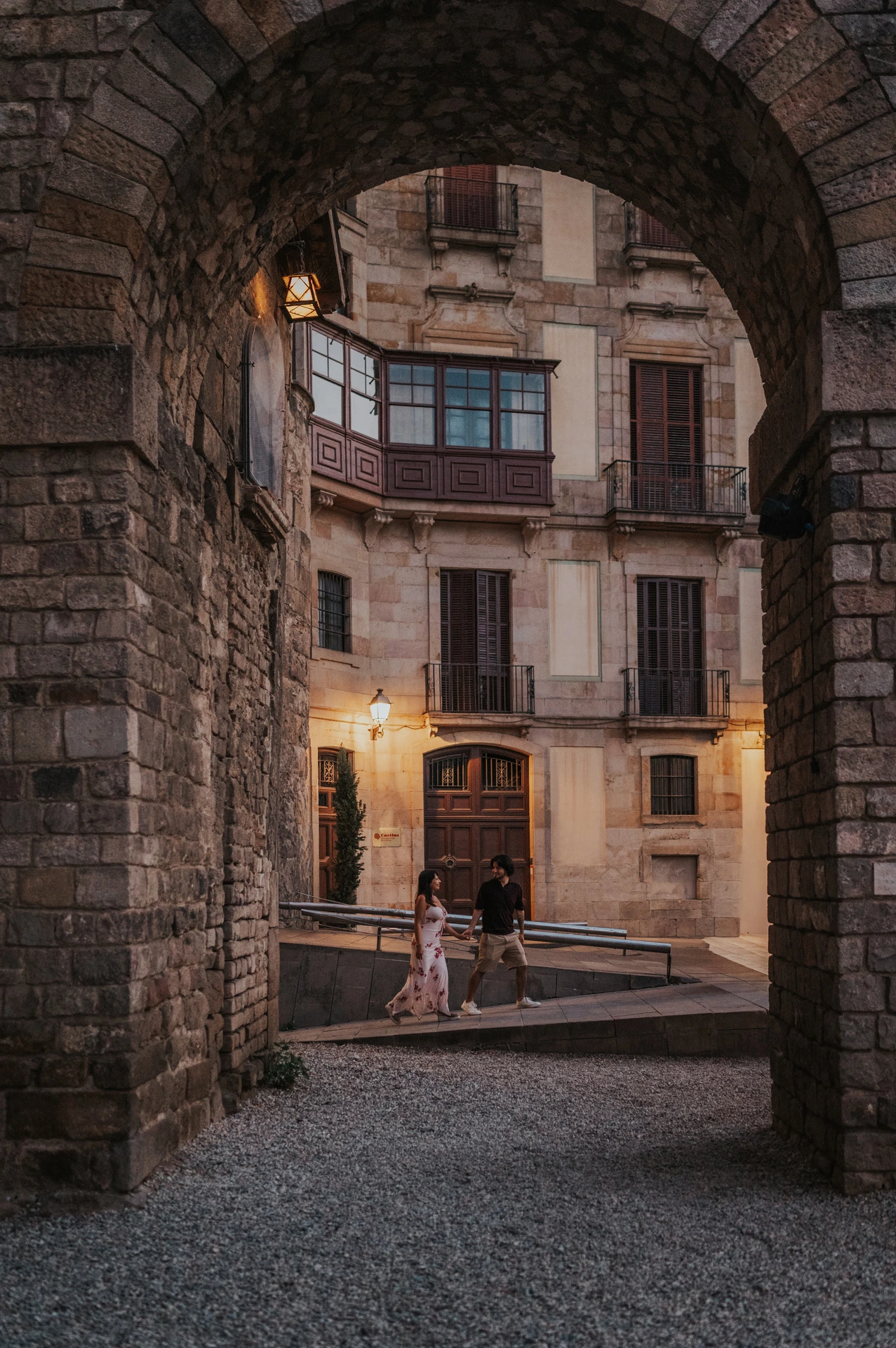 The couple walking hand in hand beneath a broad stone arch, with glowing wall lamps and old-town façades beyond them during their couple photoshoot in Barcelona.