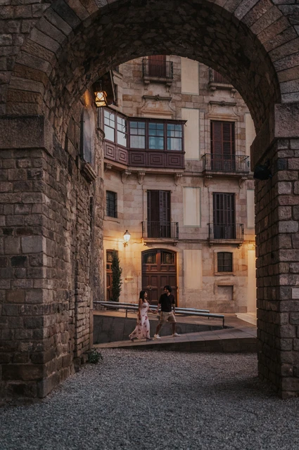 The couple walking hand in hand beneath a broad stone arch, with glowing wall lamps and old-town façades beyond them during their couple photoshoot in Barcelona.