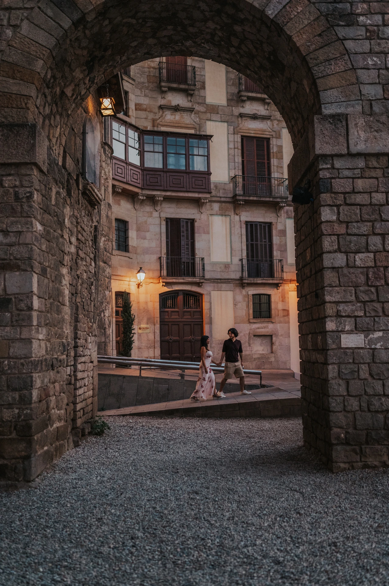 The couple walking hand in hand through a broad stone arch in the Gothic Quarter, looking towards one another during their couple photoshoot in Barcelona.