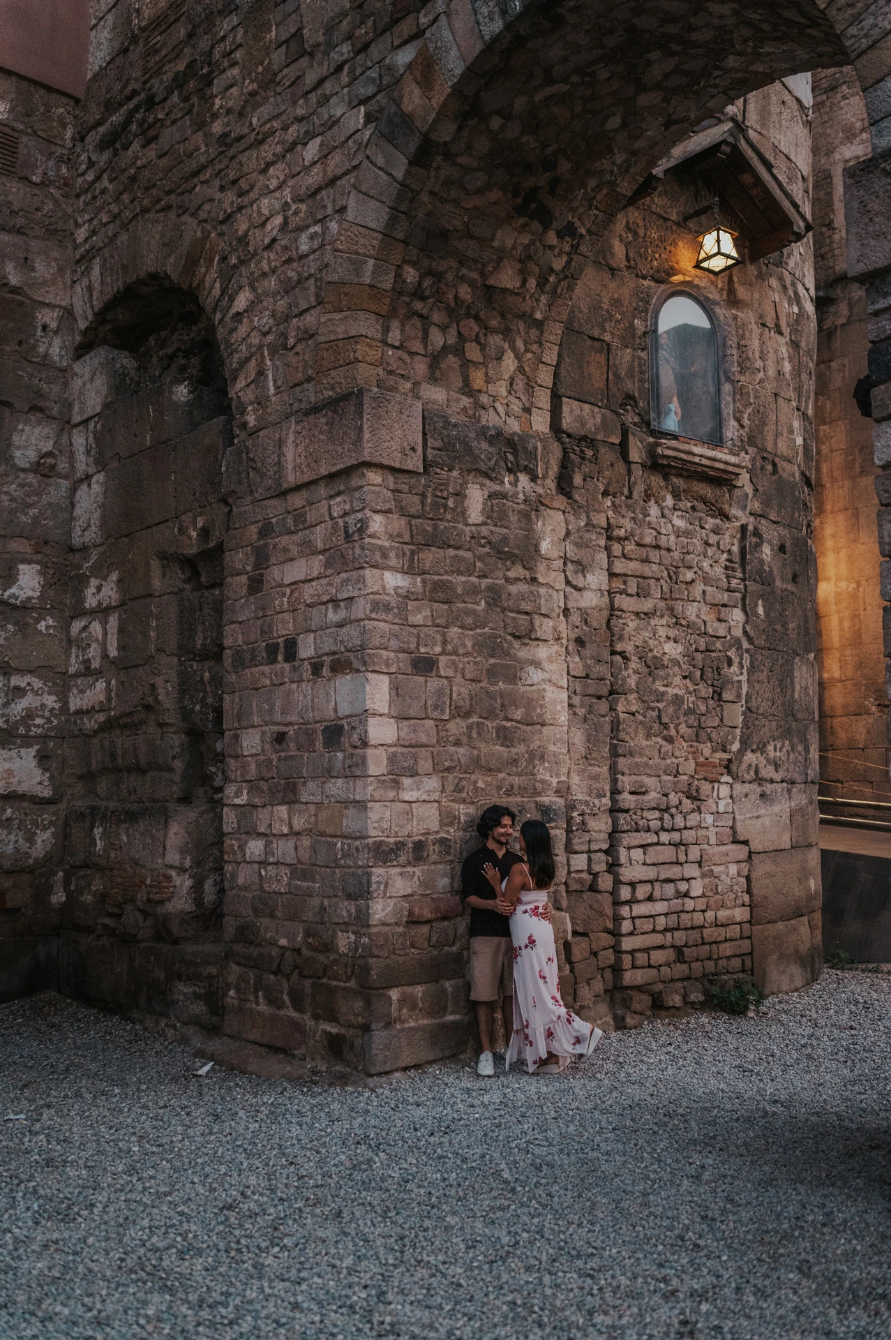 The couple standing close together at the corner of an old stone building beneath a lit wall lamp in the Gothic Quarter during their couple photoshoot in Barcelona.