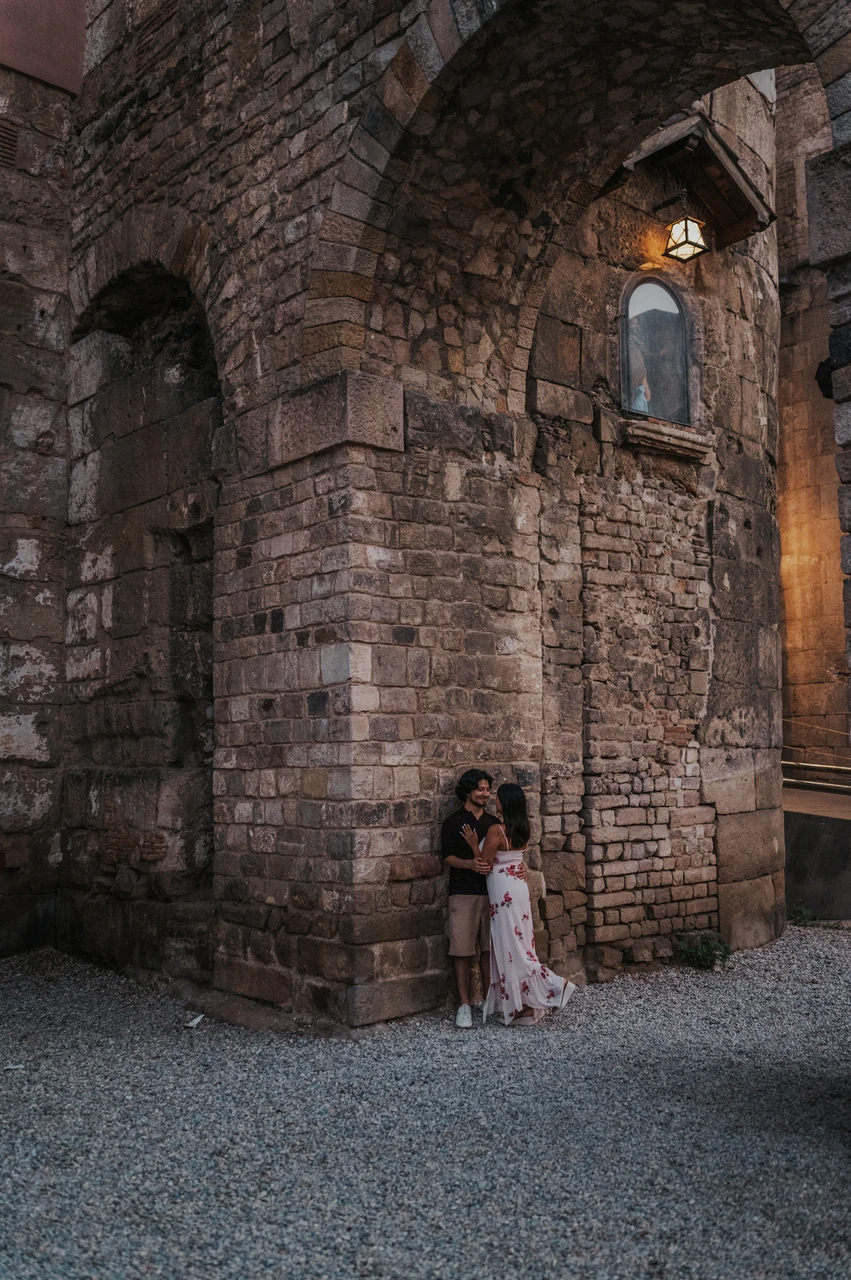 A portrait beneath a lamp at an old stone corner | Barcelona couple photoshoot The couple standing close together at the corner of an old stone building beneath a lit wall lamp in the Gothic Quarter during their couple photoshoot in Barcelona.