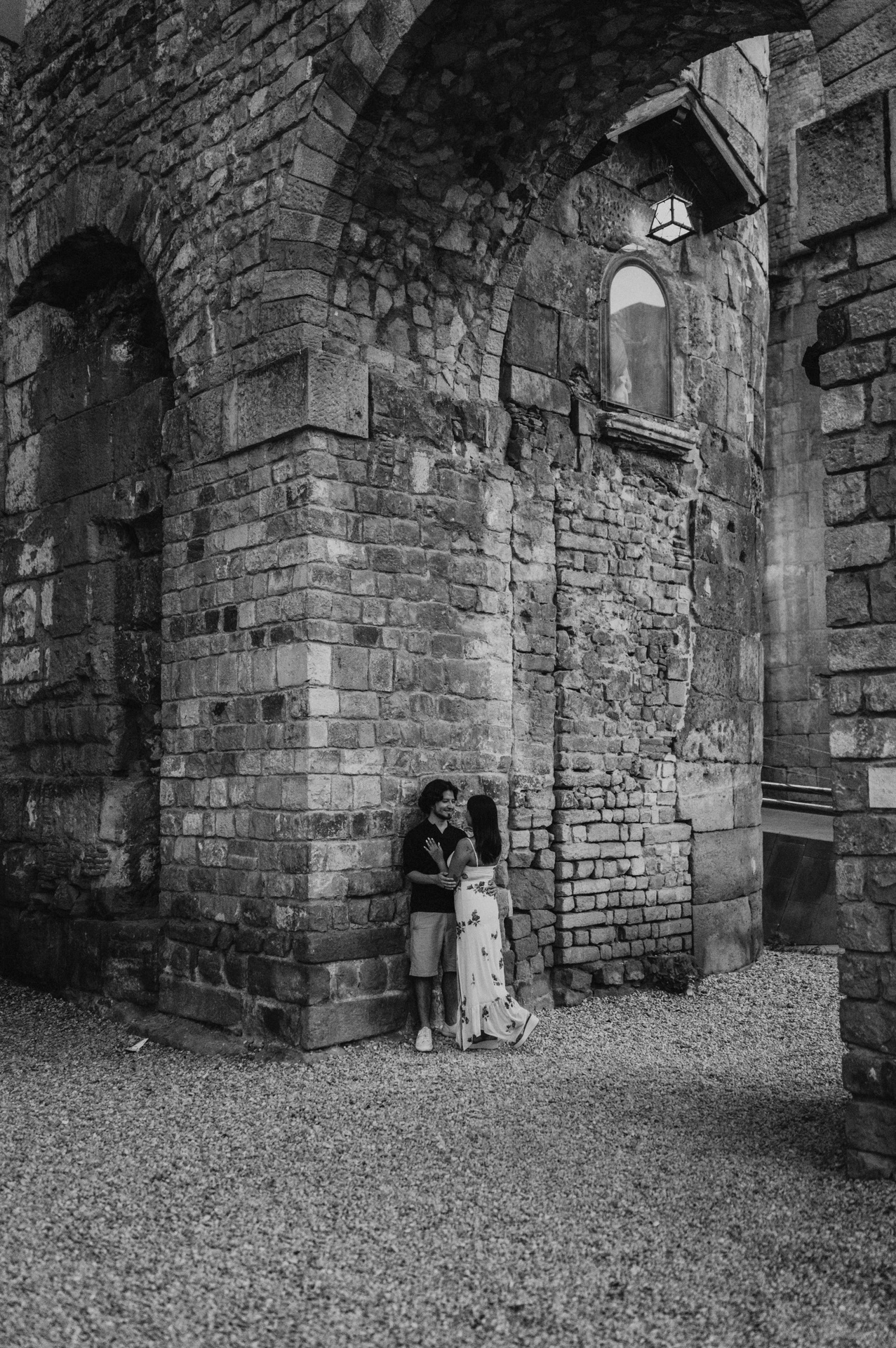 Black-and-white photograph: the couple standing close together at the corner of an old stone building beneath a lit wall lamp in the Gothic Quarter during their couple photoshoot in Barcelona.