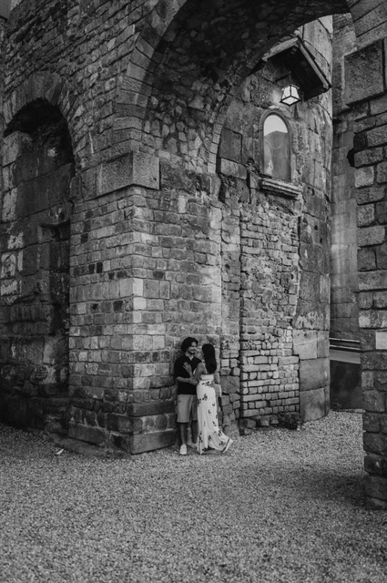 A portrait beneath a lamp at an old stone corner in black and white | Barcelona couple photoshoot Black-and-white photograph: the couple standing close together at the corner of an old stone building beneath a lit wall lamp in the Gothic Quarter during their couple photoshoot in Barcelona.
