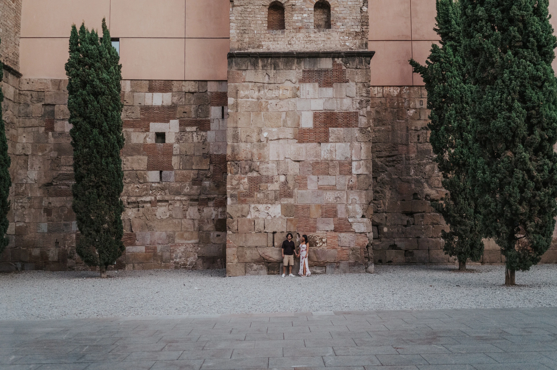A wide view of the couple beside a tall historic stone wall with cypress trees in the Gothic Quarter during their couple photoshoot in Barcelona.