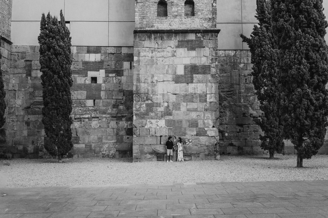 Old stone walls and cypress trees around them in black and white | Barcelona couple photoshoot Black-and-white photograph: a wide view of the couple beside a tall historic stone wall with cypress trees in the Gothic Quarter during their couple photoshoot in Barcelona.