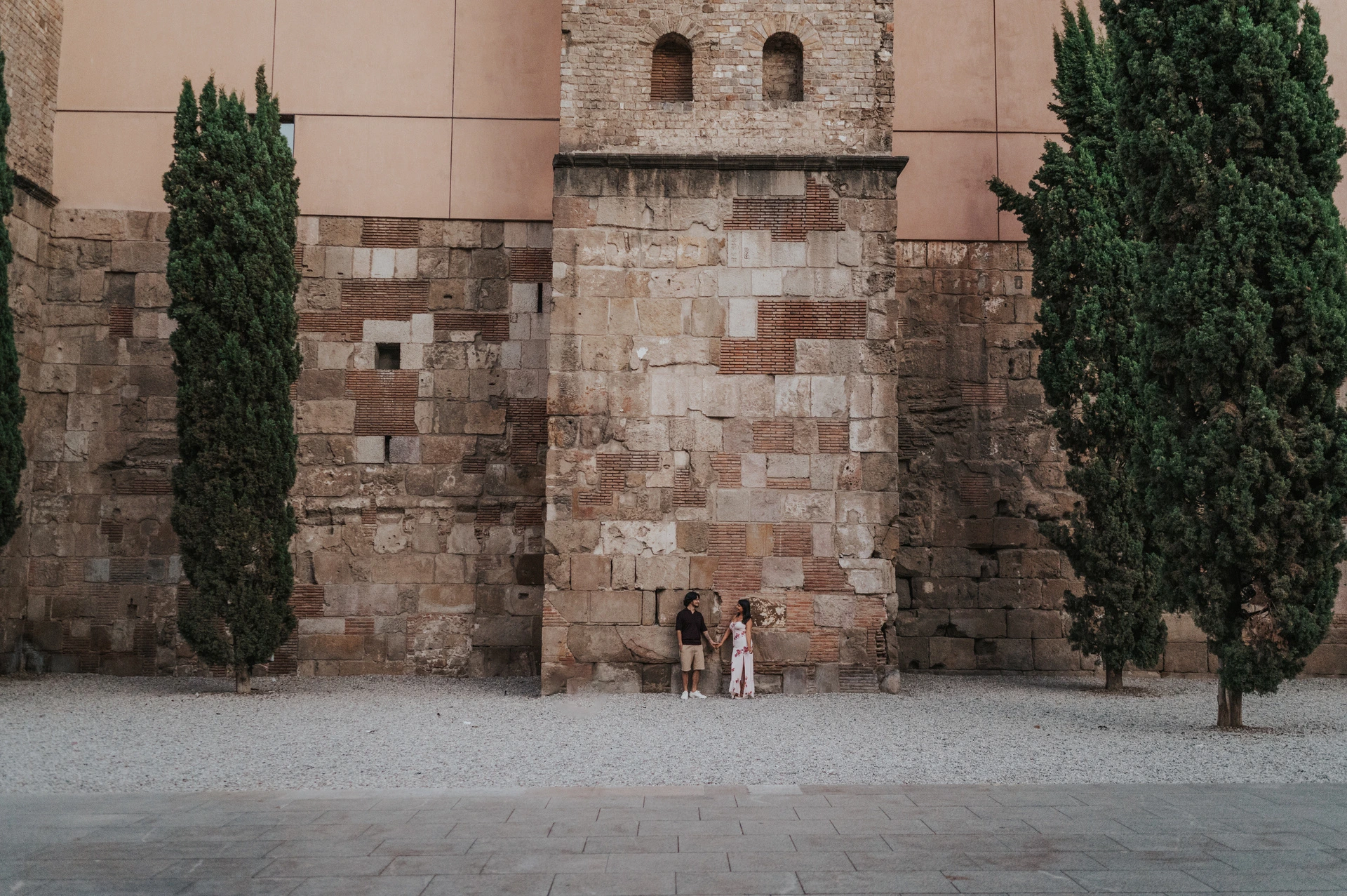 Together beneath a tall historic stone wall | Barcelona couple photoshoot The couple standing close together beside a tall historic stone wall, with cypress trees on either side in the Gothic Quarter during their couple photoshoot in Barcelona.