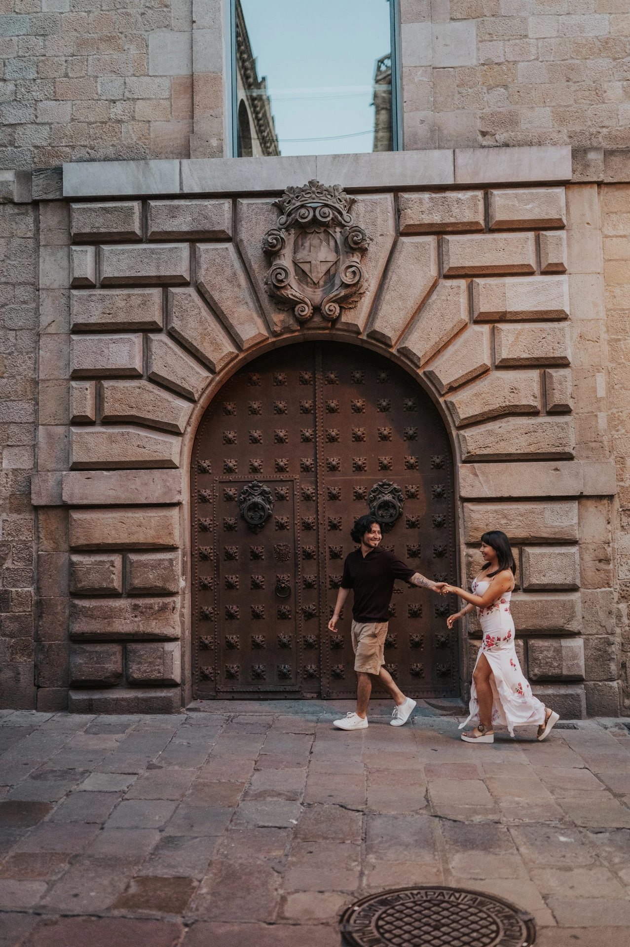 The couple facing one another and holding hands in front of a studded arched door with a carved stone face above it in the Gothic Quarter during their couple photoshoot in Barcelona.