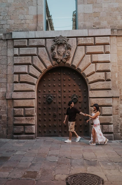 Hand in hand by a studded arched doorway | Barcelona couple photoshoot The couple facing one another and holding hands in front of a studded arched door with a carved stone face above it in the Gothic Quarter during their couple photoshoot in Barcelona.