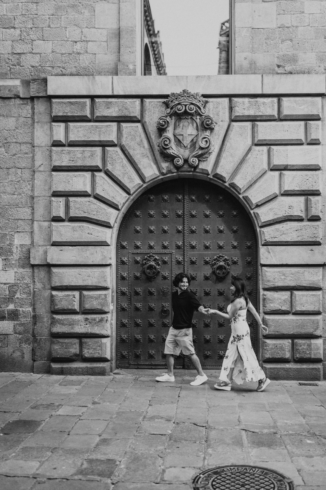 Black-and-white photograph: the couple facing one another and holding hands in front of a studded arched door with a carved stone face above it in the Gothic Quarter during their couple photoshoot in Barcelona.