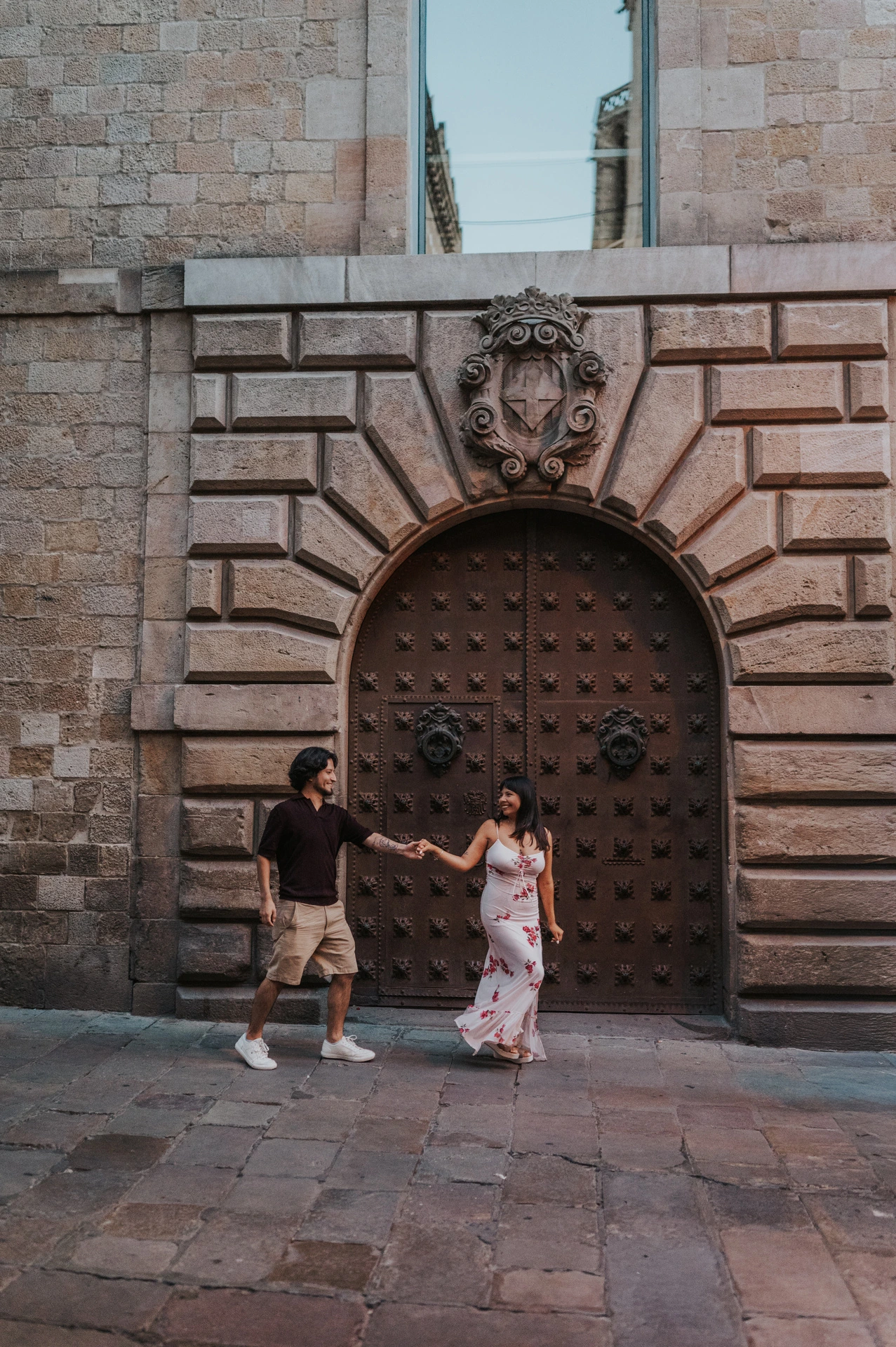 The couple walking hand in hand past a studded arched wooden door with a carved stone face above it in the Gothic Quarter during their couple photoshoot in Barcelona.