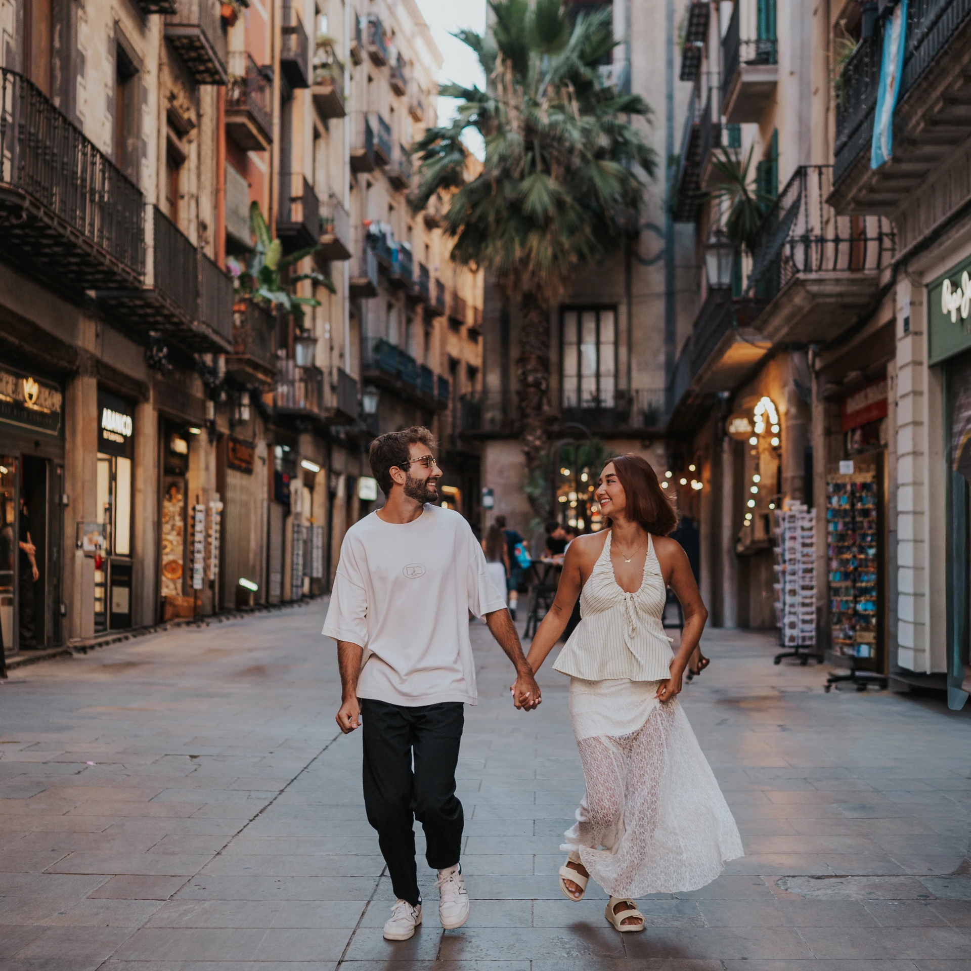 Barcelona Photoshoot Duration - El Born Walk Two adults walk hand in hand along an El Born street during a relaxed Barcelona photoshoot, framed by balconies and palms.