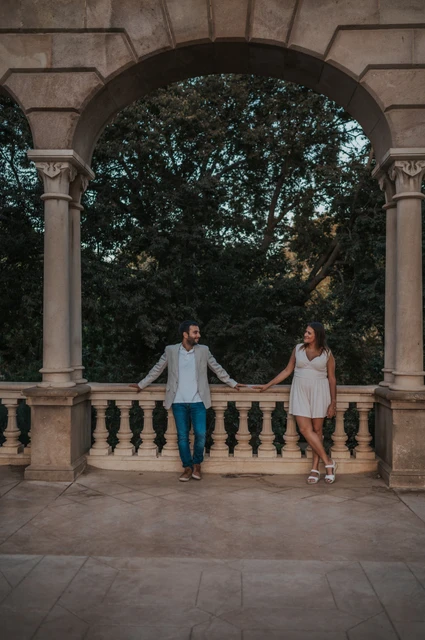 Together beneath a broad stone arch | Barcelona engagement photoshoot The couple holding hands beneath a wide stone arch, with a balustrade and trees behind them during their engagement photoshoot in Barcelona.