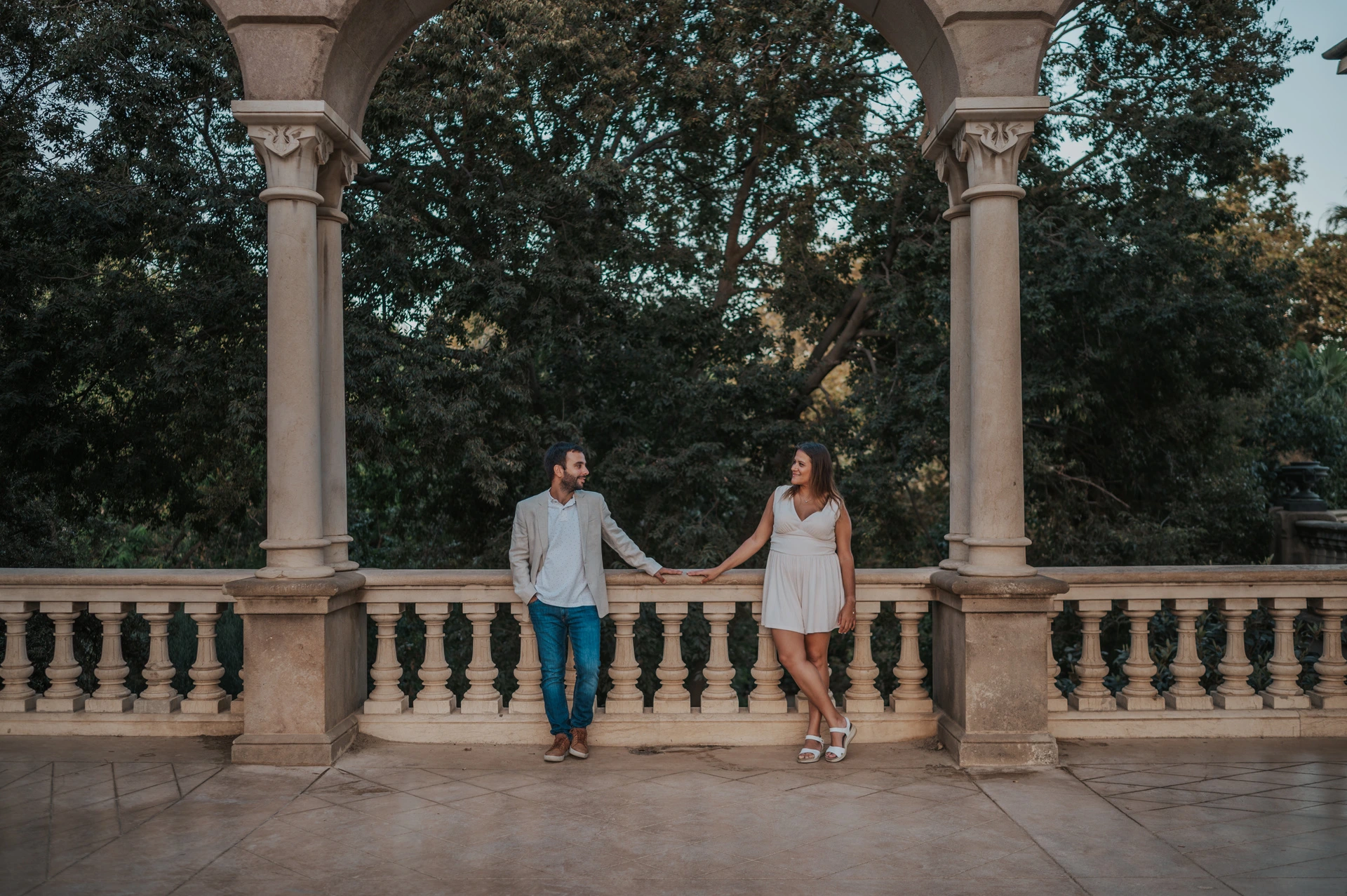 Together beneath a broad stone arch | Barcelona engagement photoshoot The couple holding hands beneath a wide stone arch, with a balustrade and trees behind them during their engagement photoshoot in Barcelona.
