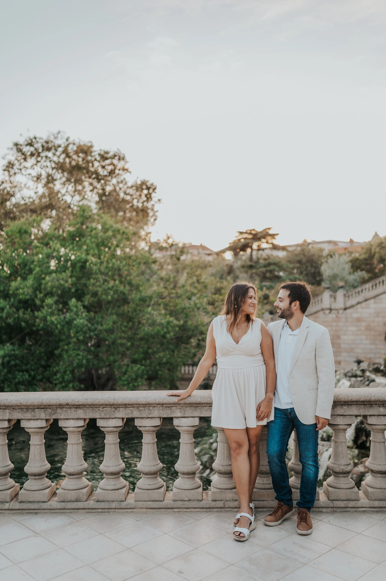 The couple standing beside a carved stone balustrade and holding hands with trees behind them during their engagement photoshoot in Barcelona.