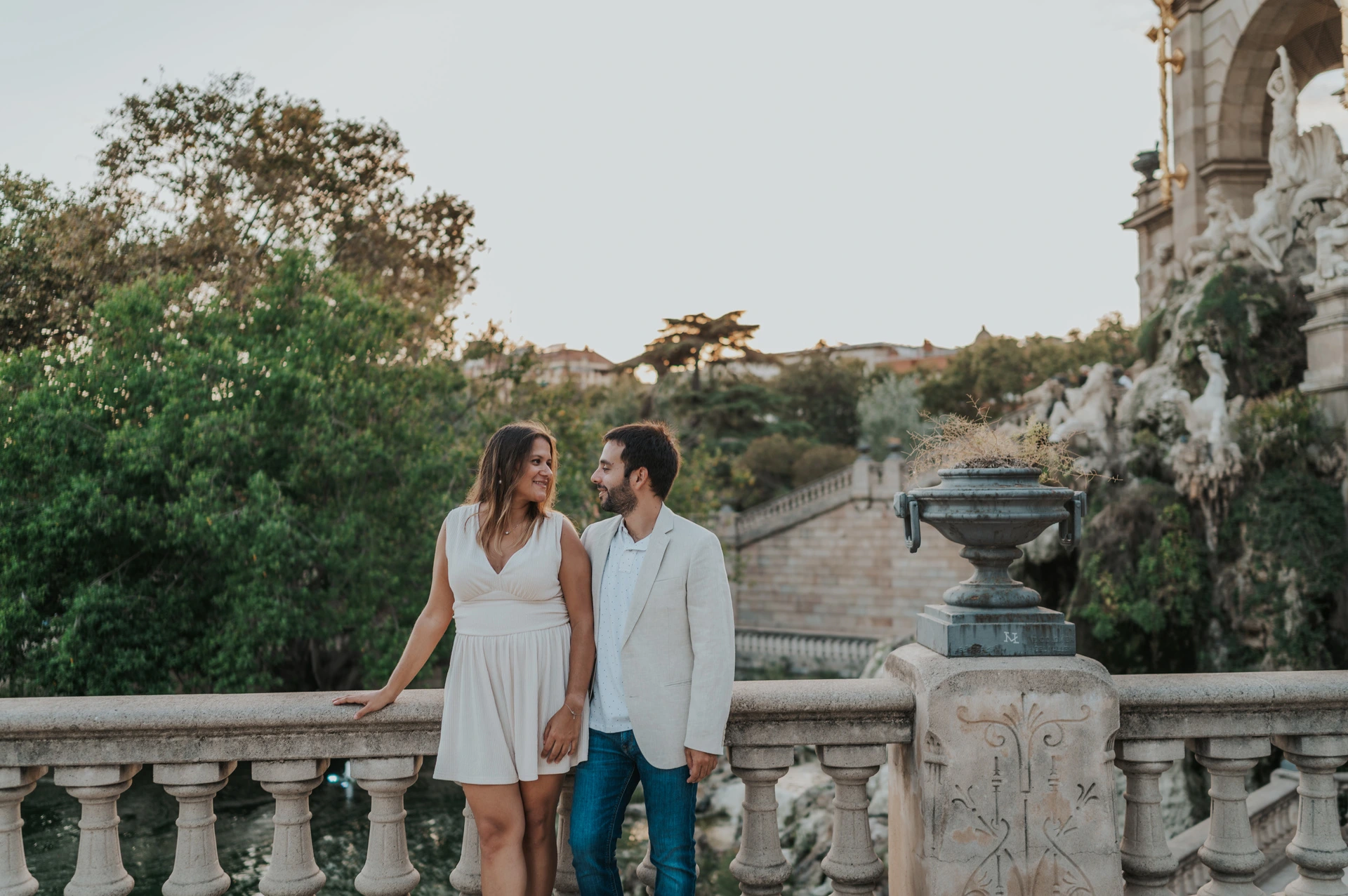 The couple standing beside a carved stone balustrade and holding hands with trees behind them during their engagement photoshoot in Barcelona.