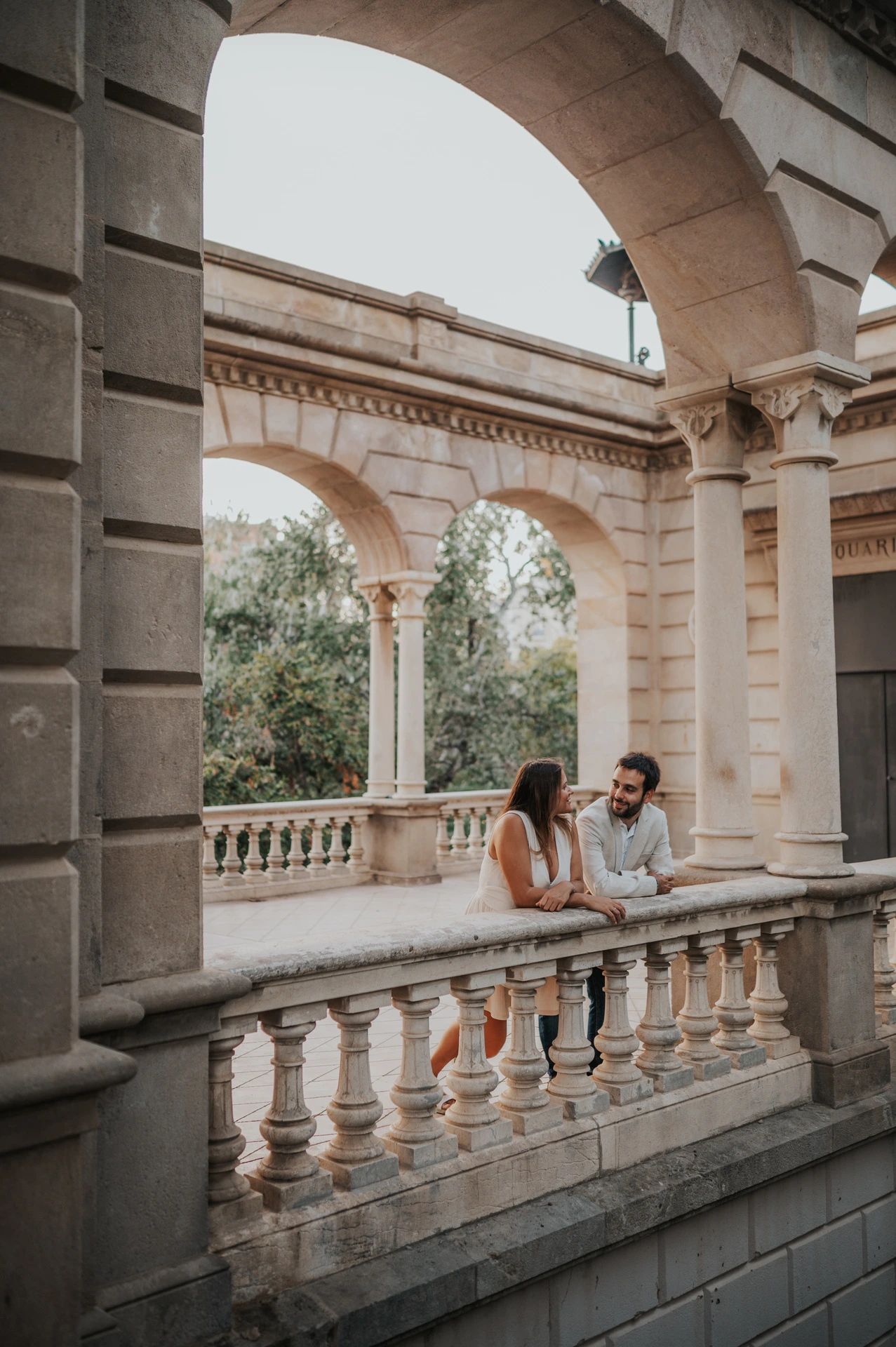 The couple leaning together on the carved stone railing beneath the pavilion arches during their engagement photoshoot in Barcelona.