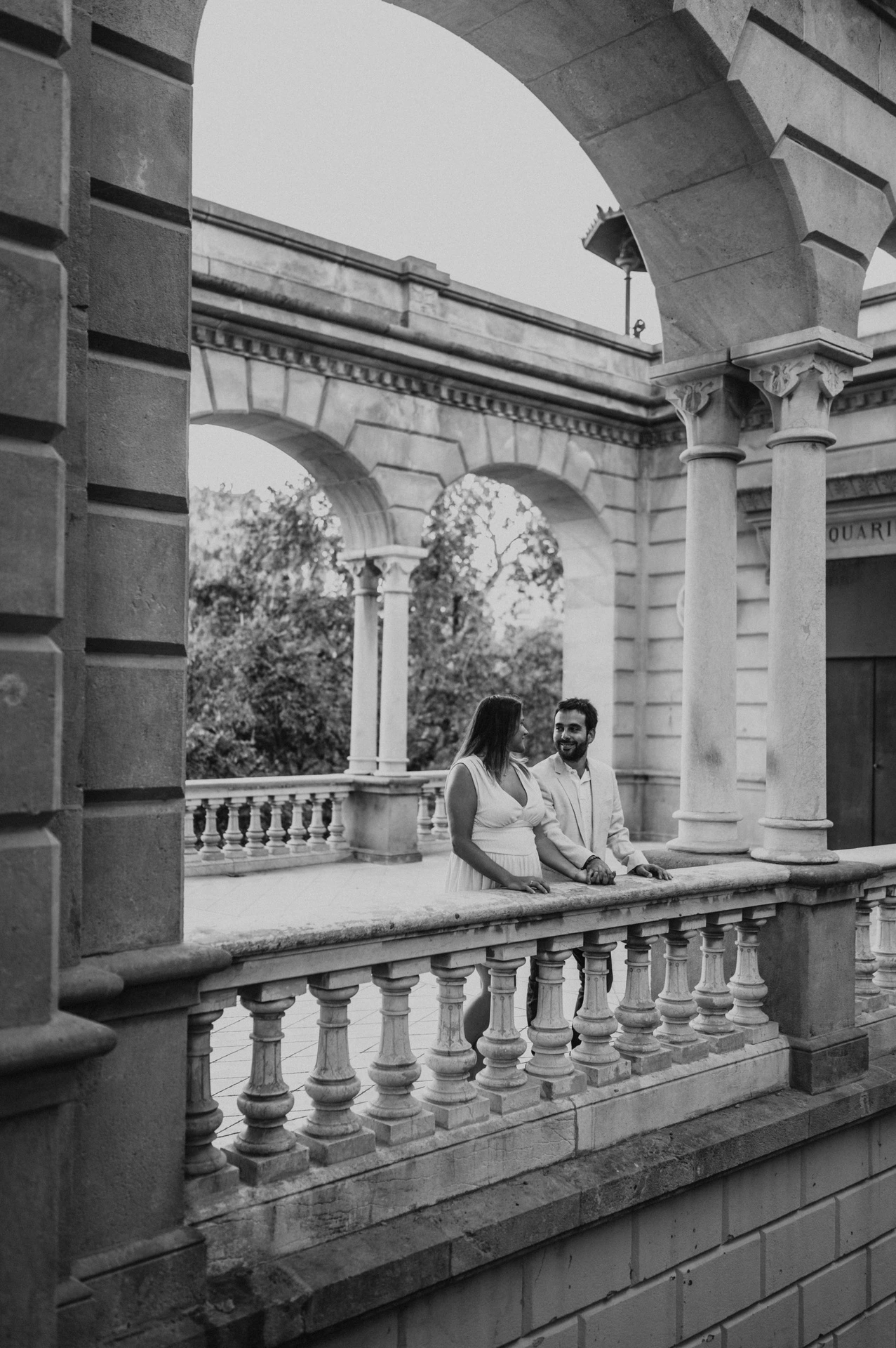 Black-and-white photograph: the couple leaning together on the carved stone railing beneath the pavilion arches during their engagement photoshoot in Barcelona.