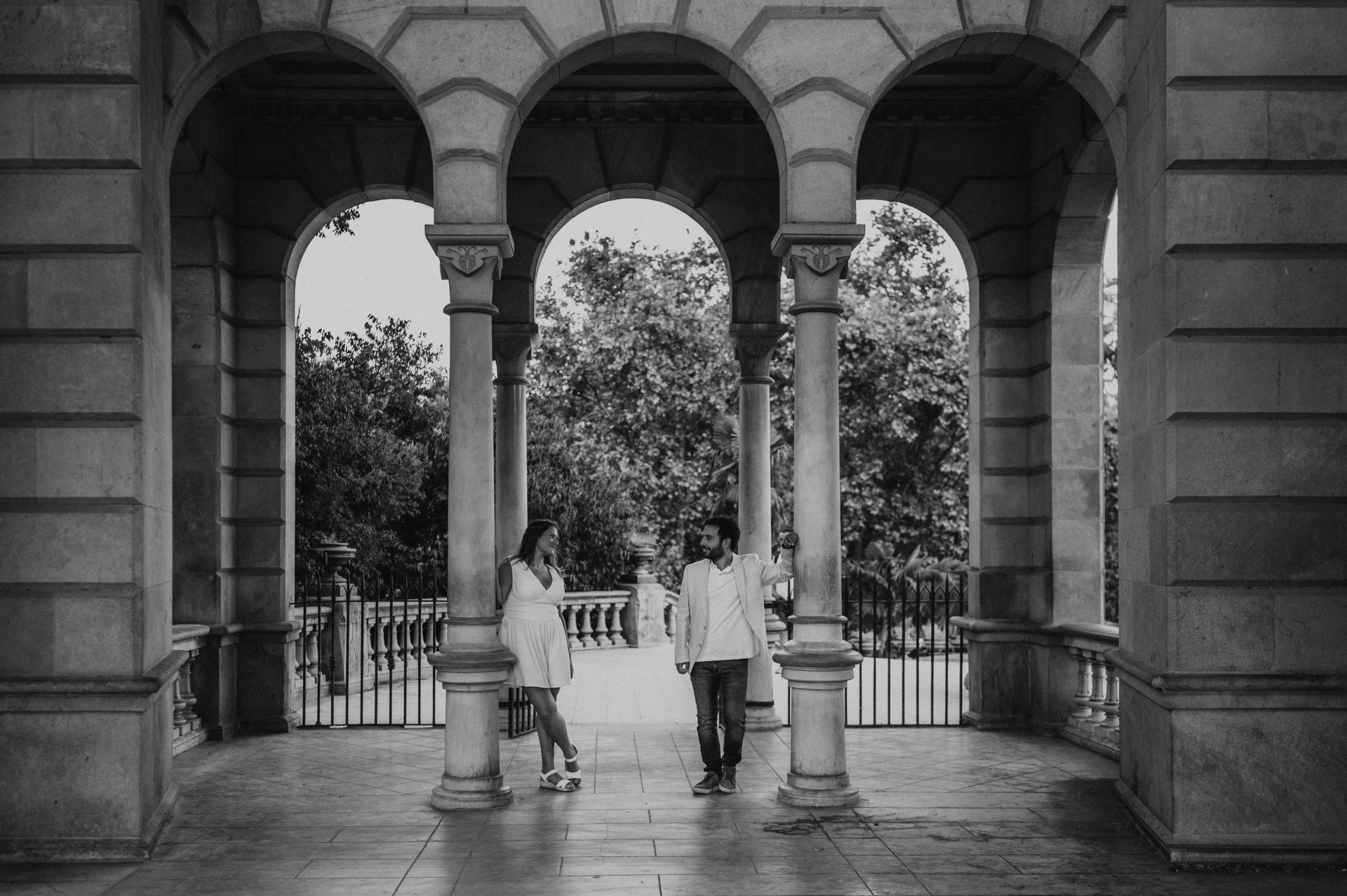Black-and-white photograph: the couple framed by the stone columns and arches of an open park pavilion during their engagement photoshoot in Barcelona.