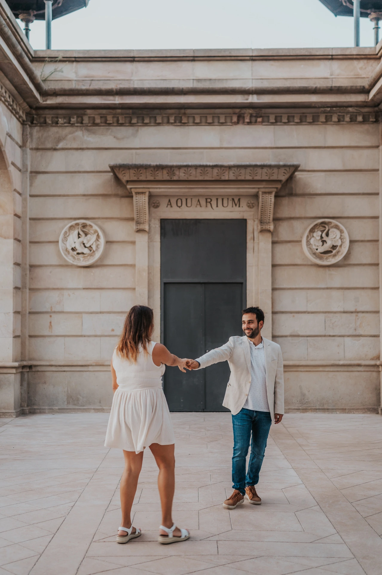 The couple holding hands as the woman turns towards the man on a paved courtyard beside the pavilion during their engagement photoshoot in Barcelona.