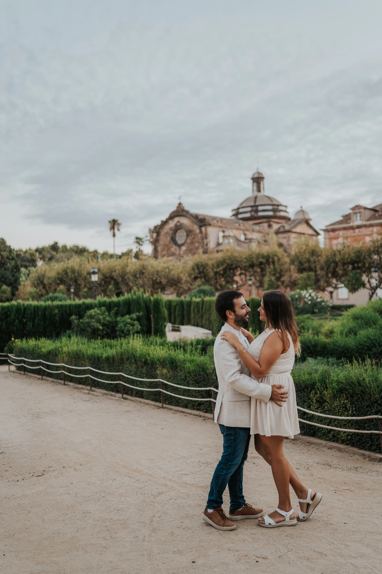 The couple kissing in front of clipped hedges and a clock-topped park building during their engagement photoshoot in Barcelona.