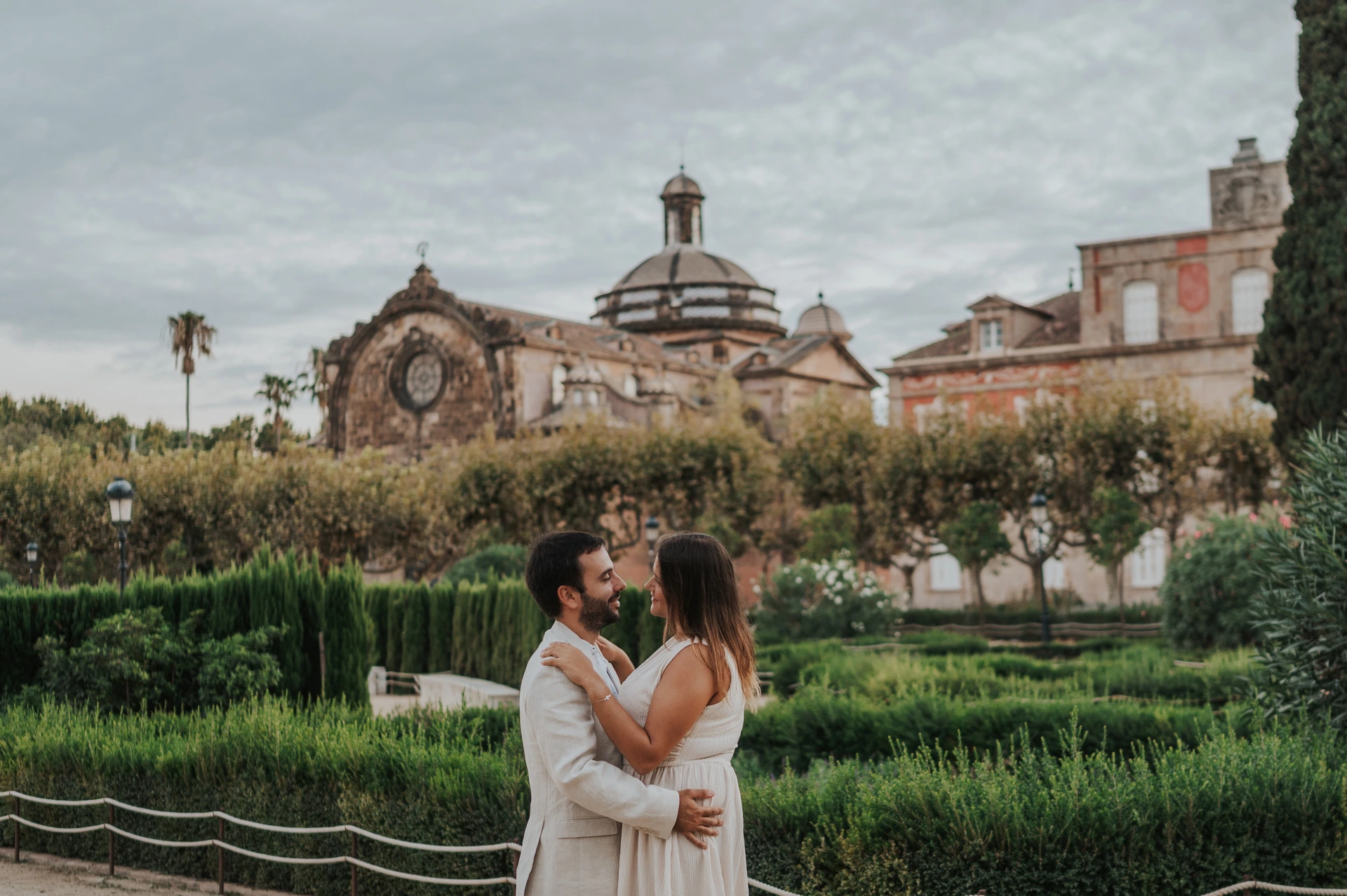 A kiss beside the formal gardens | Barcelona engagement photoshoot The couple kissing in front of clipped hedges and a clock-topped park building during their engagement photoshoot in Barcelona.