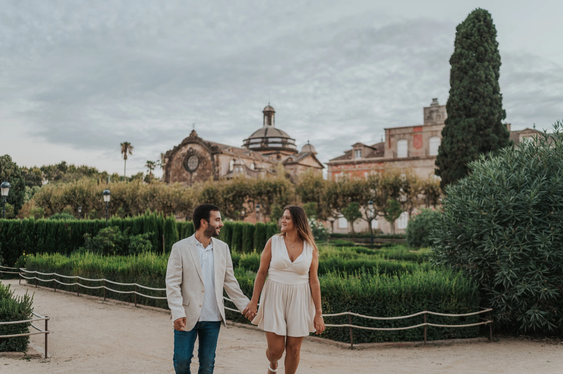 The couple walking hand in hand along a path beside clipped hedges and a clock-topped building during their engagement photoshoot in Barcelona.