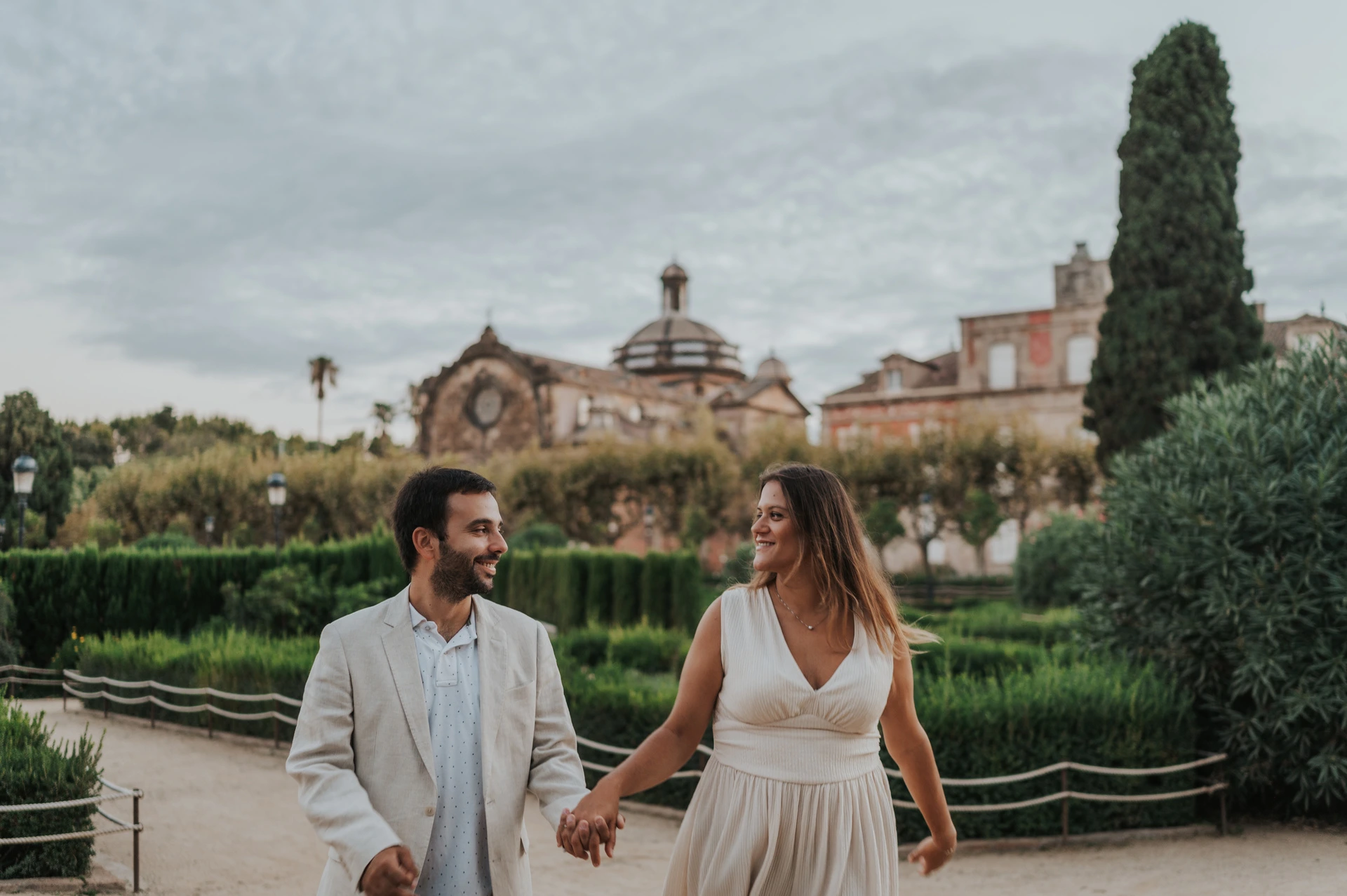The couple walking hand in hand along a path beside clipped hedges and a clock-topped building during their engagement photoshoot in Barcelona.