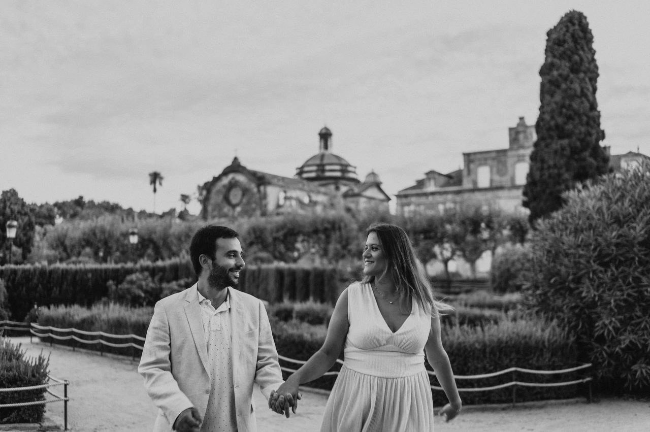A hand-in-hand walk beside the garden hedges in black and white | Barcelona engagement photoshoot Black-and-white photograph: the couple walking hand in hand along a path beside clipped hedges and a clock-topped building during their engagement photoshoot in Barcelona.
