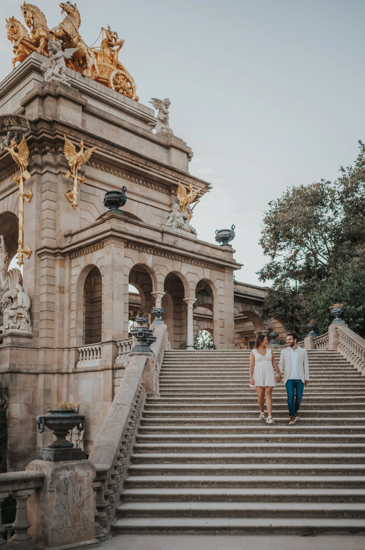 The couple walking down the broad stone staircase beside the fountain monument during their engagement photoshoot in Barcelona.