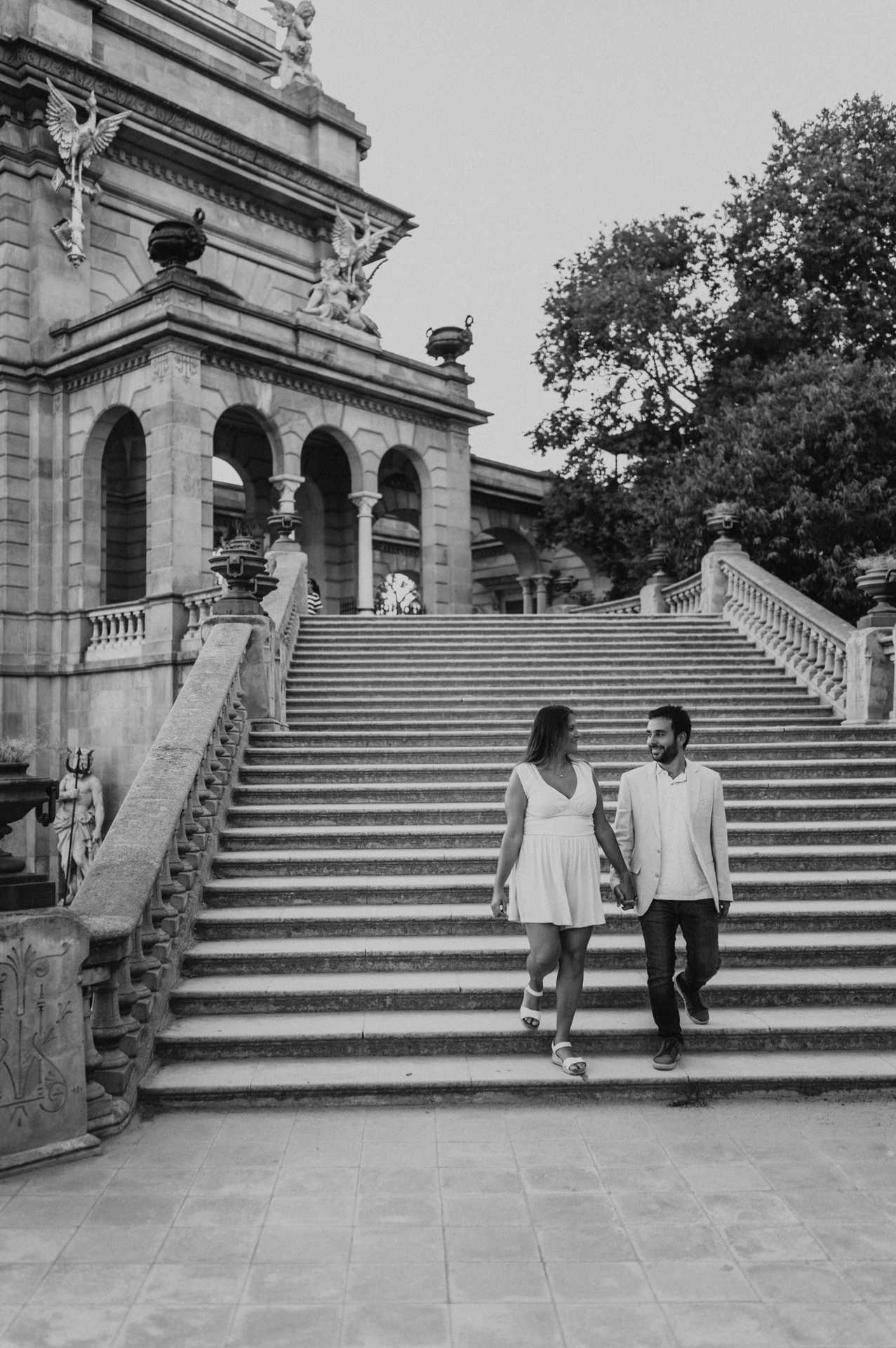Black-and-white photograph: the couple walking down the broad stone staircase beside the fountain monument during their engagement photoshoot in Barcelona.