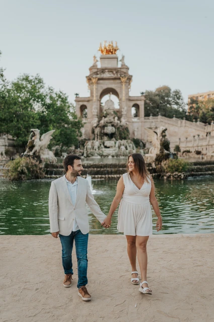 The couple walking hand in hand beside the green fountain pool, with the ornate monument behind them during their engagement photoshoot in Barcelona.