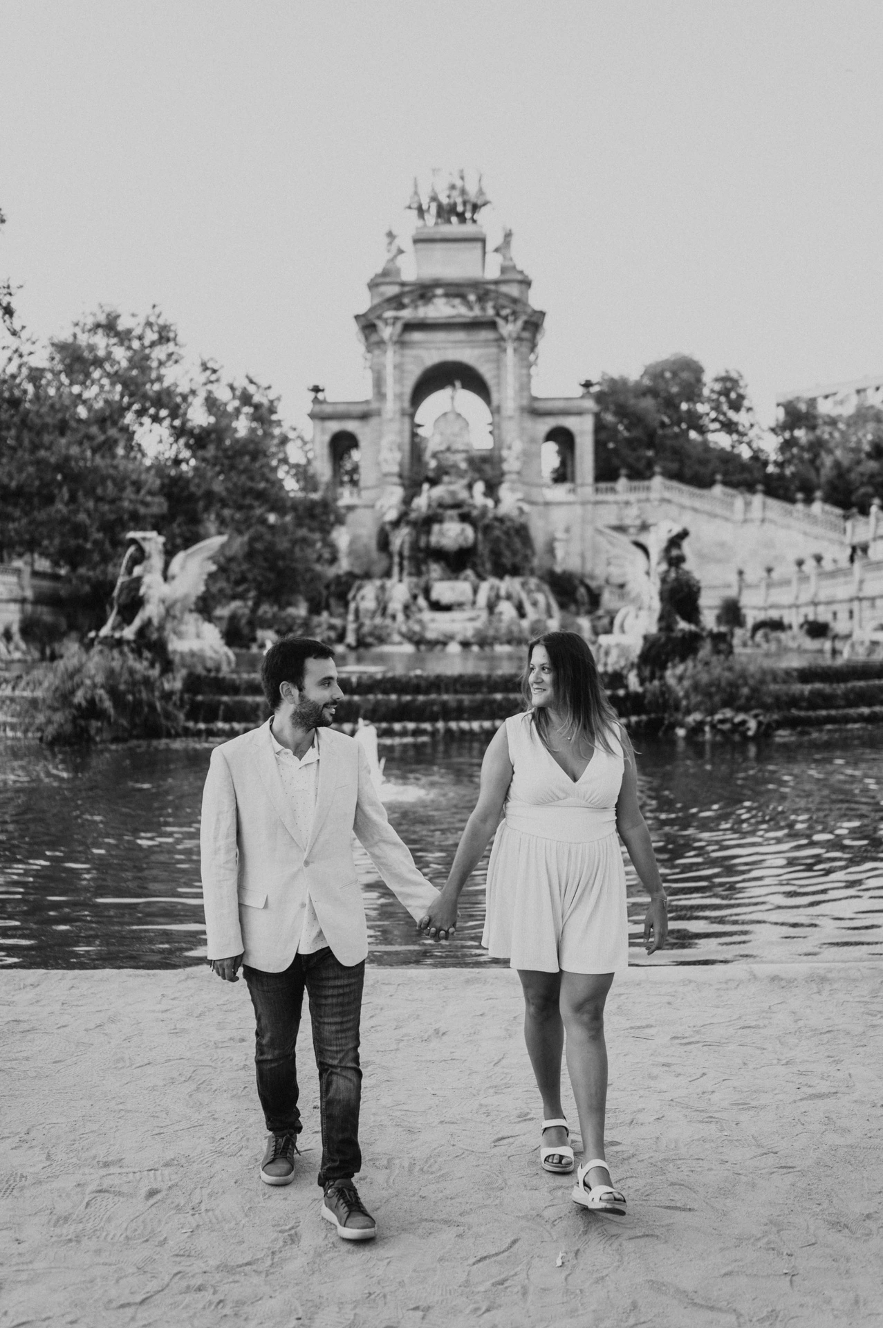 Black-and-white photograph: the couple walking hand in hand beside the green fountain pool, with the ornate monument behind them during their engagement photoshoot in Barcelona.