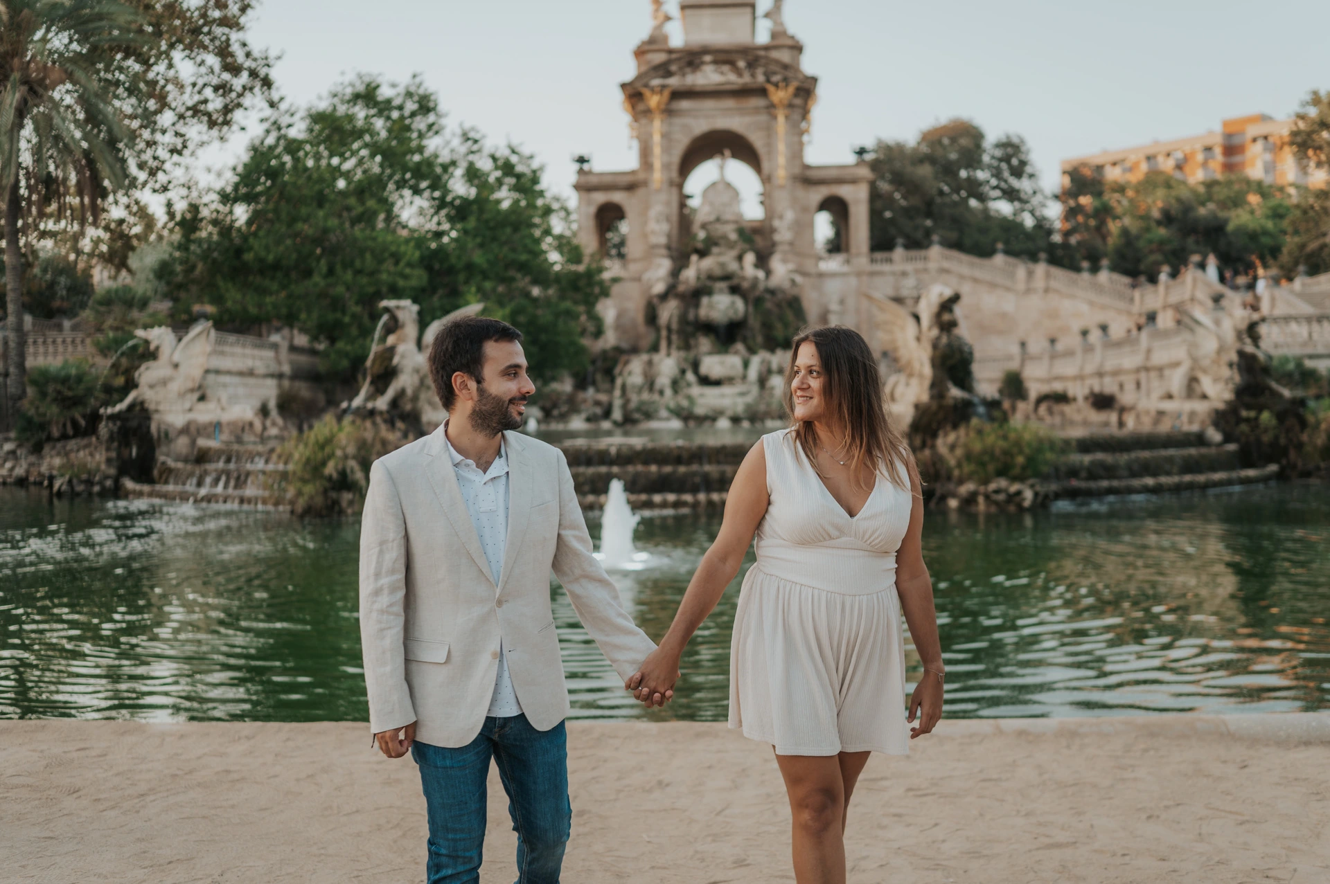 A hand-in-hand walk beside the fountain | Barcelona engagement photoshoot The couple walking hand in hand beside the green fountain pool, with the ornate monument behind them during their engagement photoshoot in Barcelona.